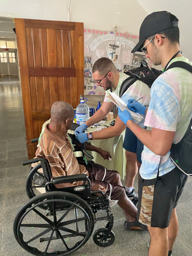 Photo of a man in a wheel chair and two student nurses, Mission Trip to Jamaica, Elms College
